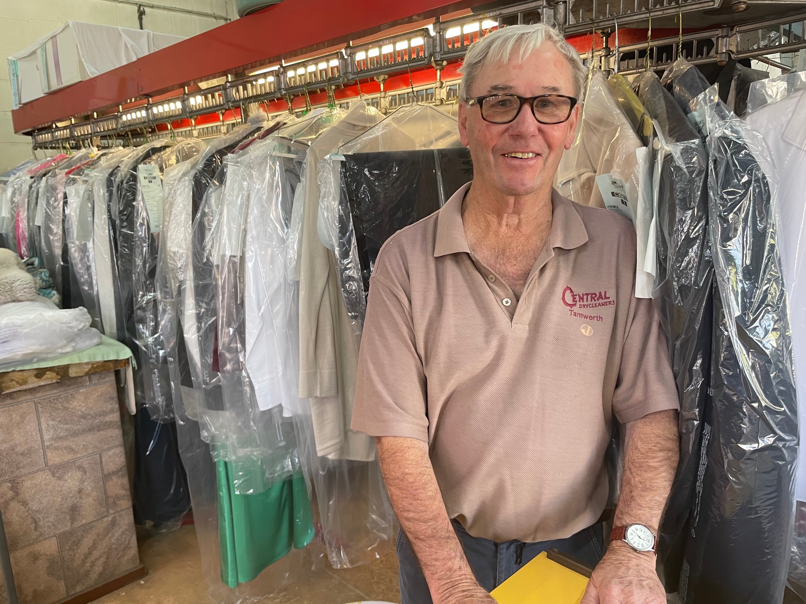 An older gentlalnd with grey hair and glasses stands in front of dozens of freshly ironed and covered clohtes on coathangers