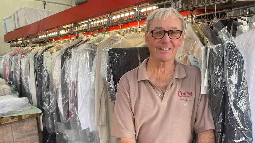 An older gentlalnd with grey hair and glasses stands in front of dozens of freshly ironed and covered clohtes on coathangers