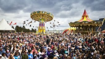 A large crowd gathers in front of show rides including a merry-go-round.