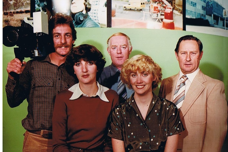 Three men, one holding an old fashioned camera, and two women standing in front of a TV studio set.