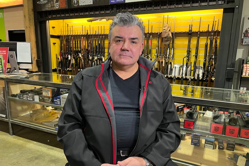 A man seating in front of a rack of rifles