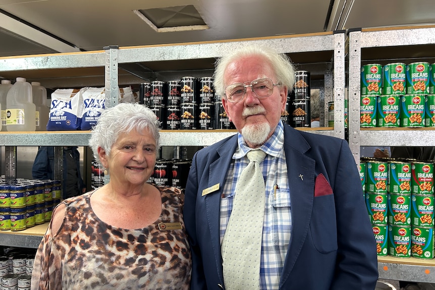 A woman in a leopard-print top with white hair and a man with glasses and a goatee stand in front of a shelf filled with cans