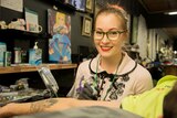Ash Chain takes a moment to smile for a photograph while tattooing a cartoon character onto the inside of a man's bicep.