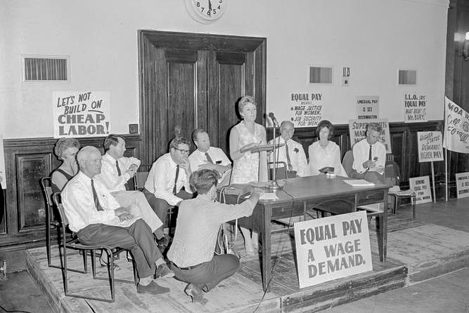 A women's rights campaigner, surrounded by men, speaks into a microphone. Placards calling for equal pay are placed around her