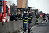 A motorway is closed as a charred bus sits up against a concrete divider with Italian police in navy and yellow hi-vis.