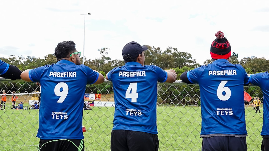 Seven men in blue soccer jerseys lean against a fence, watching a soccer match.