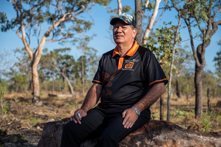 A man in a hat and branded polo shirt sits on a rock in bushland, holding a piece of cotton.
