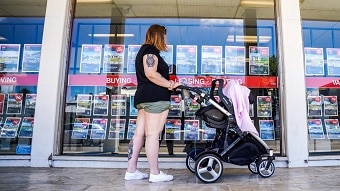 A woman stands with a pram looking at a large glass window with properties for sale pinned to it.