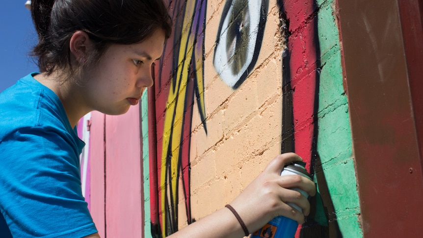 A teenage girl paints a cartoon woman on a brick wall using a spray can