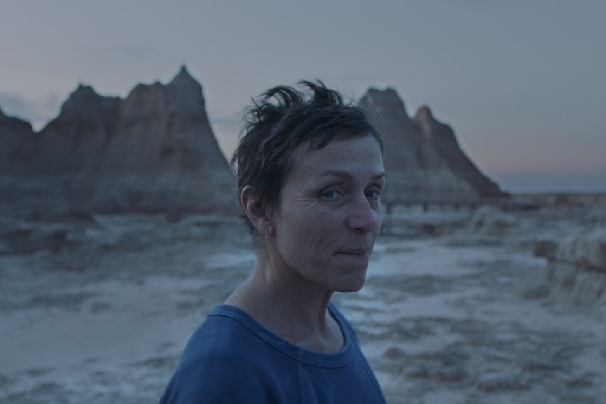 Twilight shot of the actress with pixie haircut and no make-up, wearing blue t-shirt, with arid mountain landscape in background