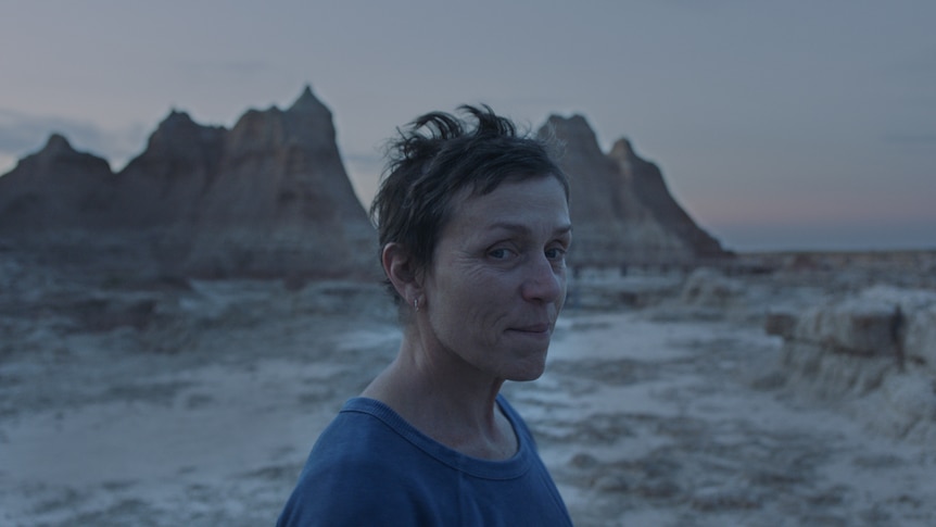 Twilight shot of the actress with pixie haircut and no make-up, wearing blue t-shirt, with arid mountain landscape in background