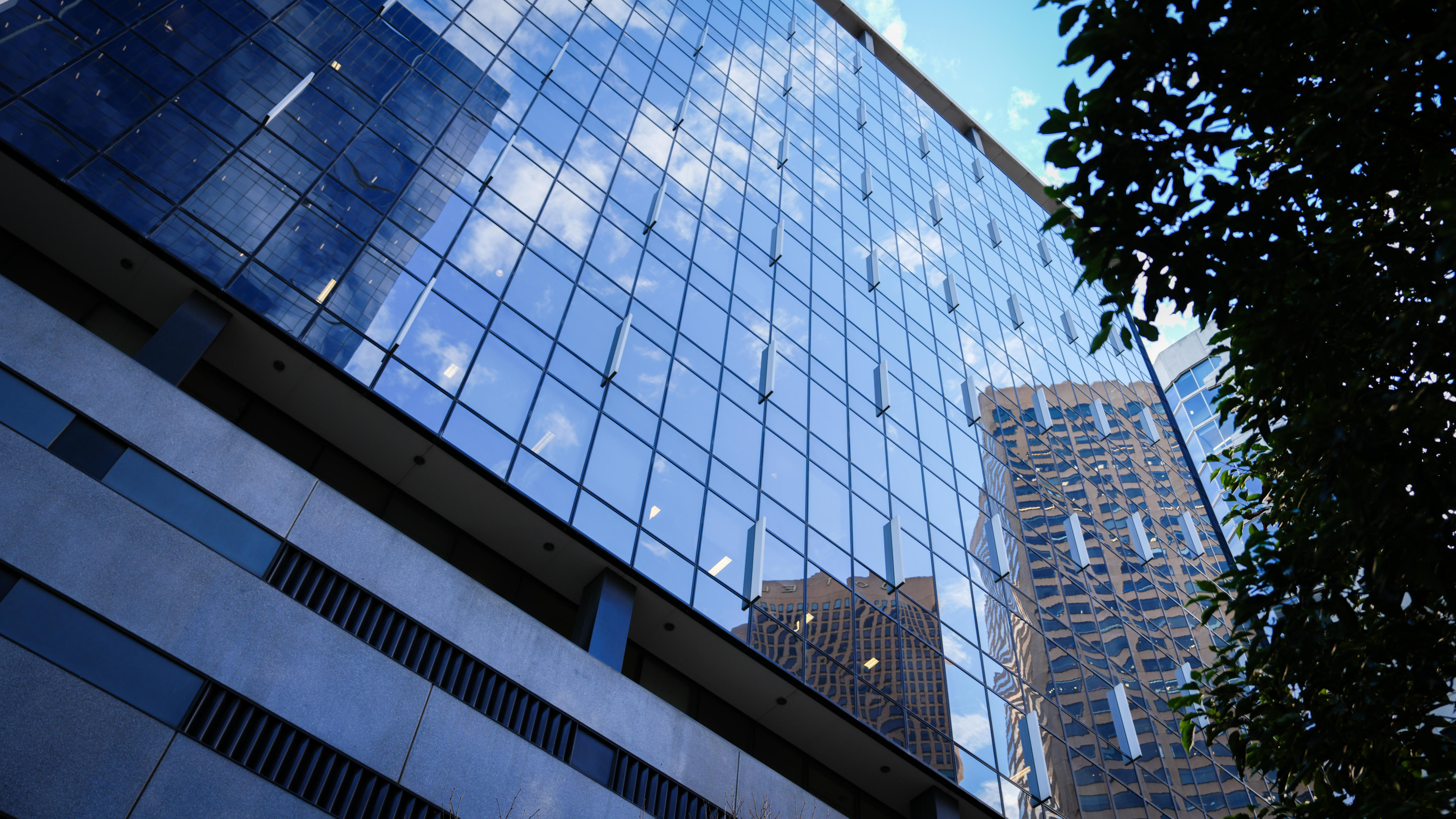 A picture taken looking up the glass facade of a multi-storey building that reflects highrises around it under a blue sky.