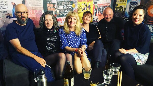 Frente! and Sally Seltmann sit on couches in a postered backstage area. On a table in front of them is a bottle and glasses.