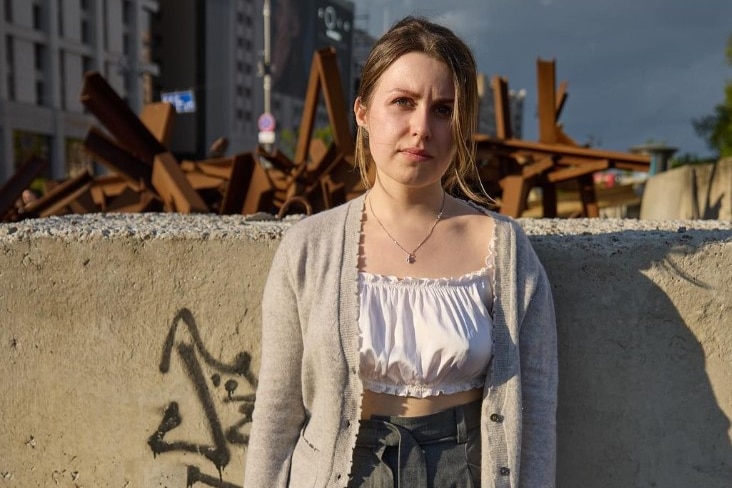A woman stands in front of a wall in Kyiv. Behind the wall there appears to be a timber baricade