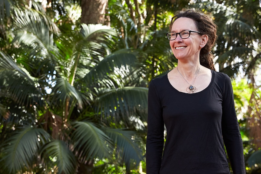 A woman standing outside with a tropical garden in the background, wearing a black long-sleeve top and black rimmed glasses. 