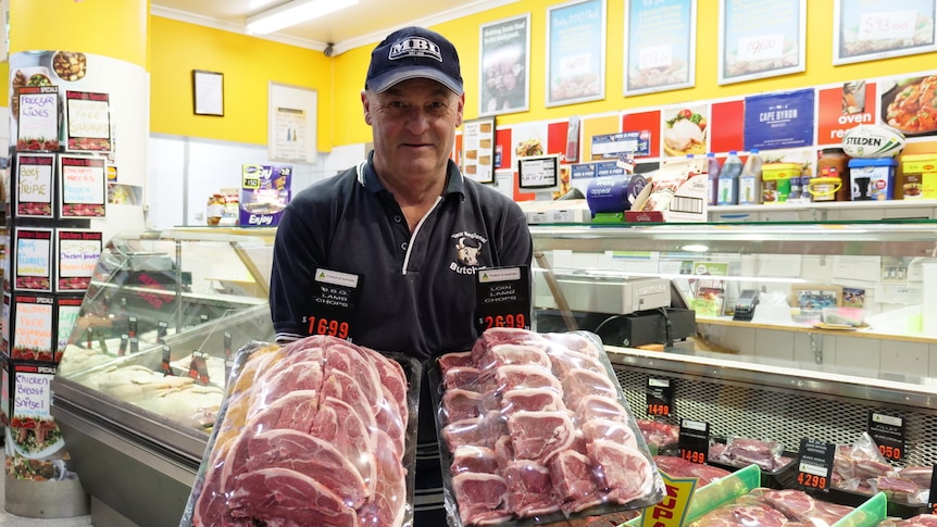 Man standing in a butcher shop holding trays of lamb.
