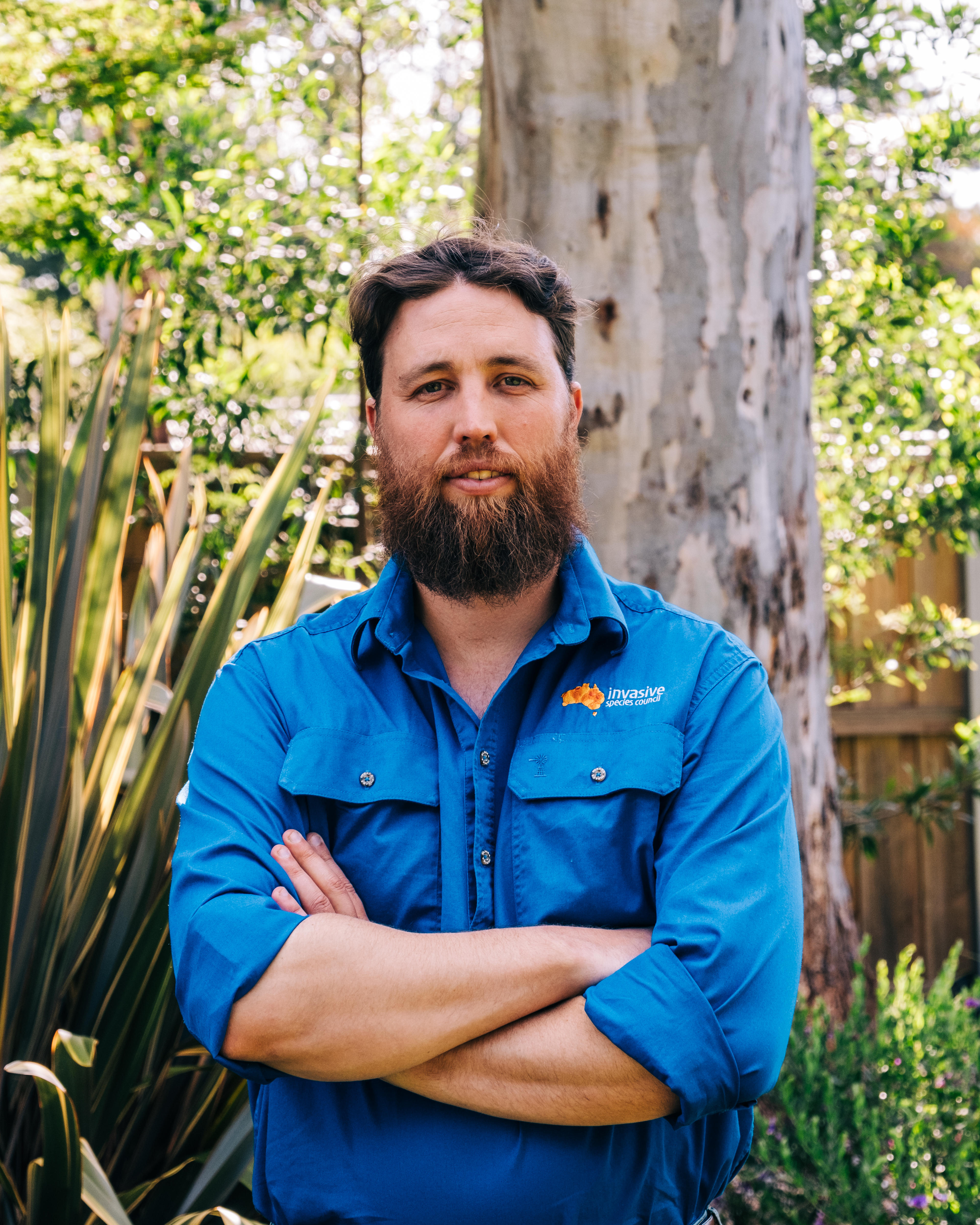 Um homem barbudo com os braços cruzados fica em frente ao tronco de uma grande árvore de eucalipto