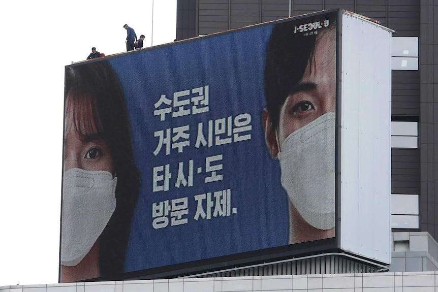 A blue billboard with face close-ups of young woman and man wearing face masks, with Korean writing.