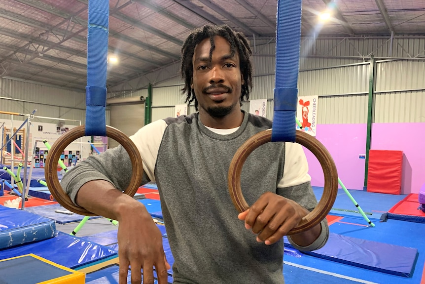 Gymnast coach Emeka Ekezie stands in a gymnastics space,  with hands leaning into gymnastic rings
