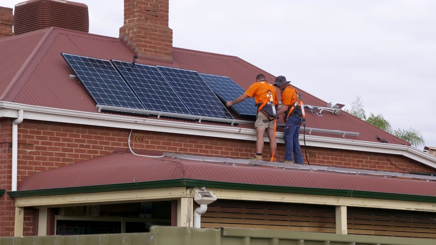 Two men install solar panels on the roof of a red-brick and red-roofed cottage