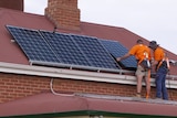 Two men install solar panels on the roof of a red-brick and red-roofed cottage