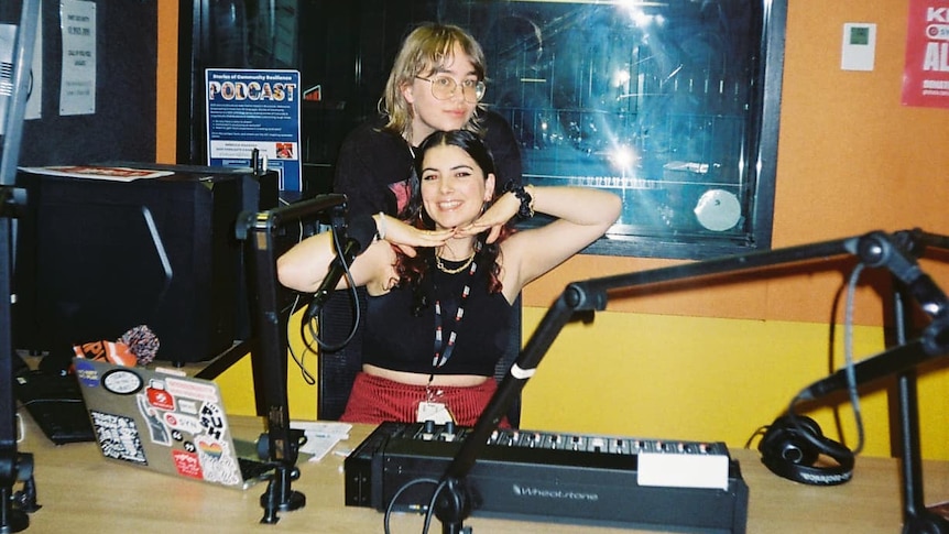 A film photo of two women embracing and smiling at a desk inside a radio studio.