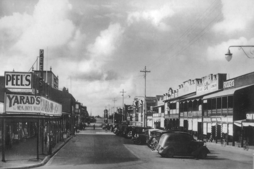 A black and white photo of businesses and vintage cars in the main street of a country town.