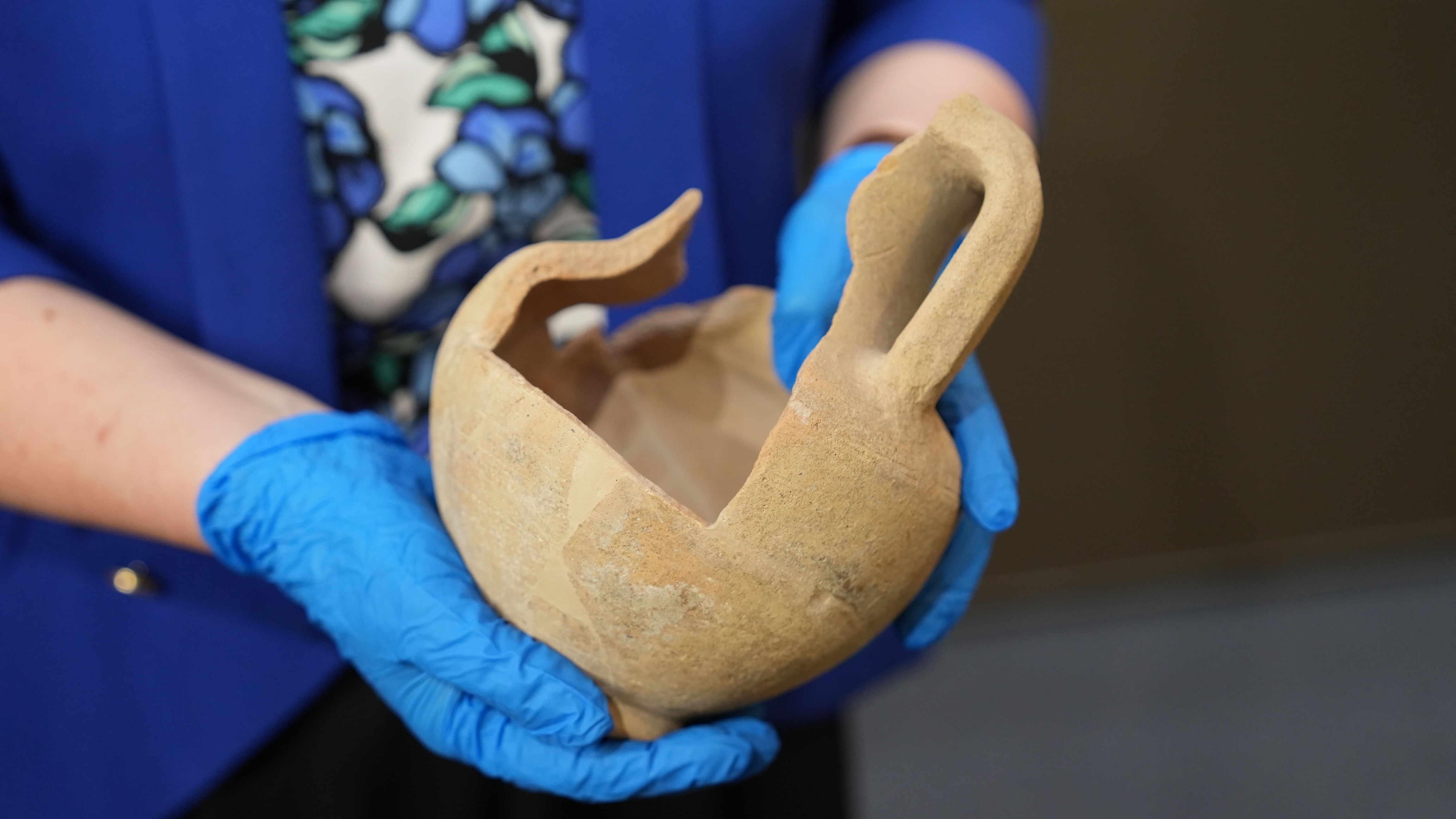 A person wearing blue latex gloves holds a partly broken ancient terracotta amphora.
