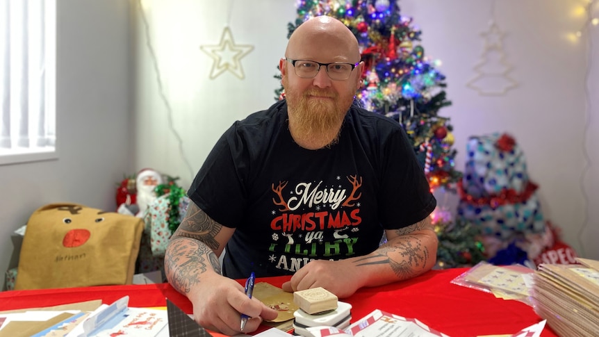 A man wearing glasses sits at a desk with letters in front of him. There is a Christmas tree in the background.