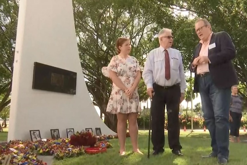 A woman and two men stand by a memorial covered in wreaths