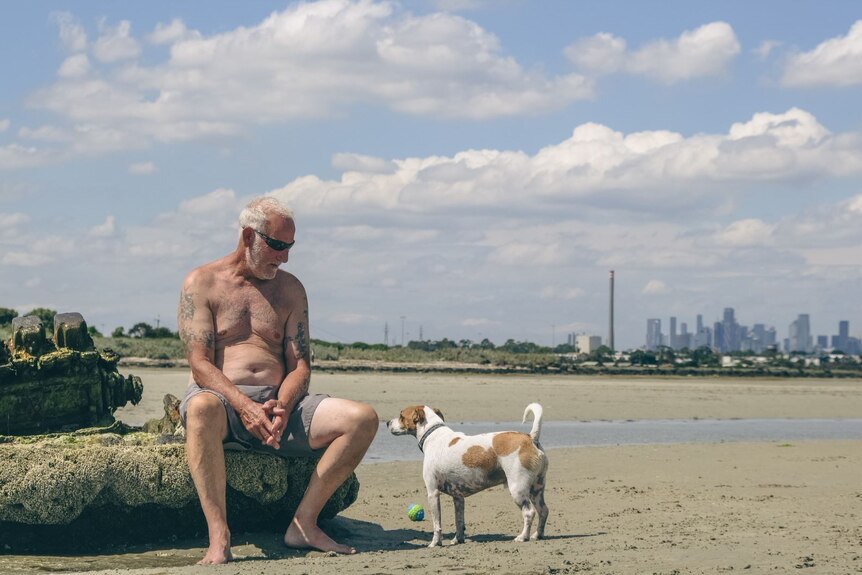 A man sits on beach debris with the city skyline behind him, and a small brown and white dog near his feet