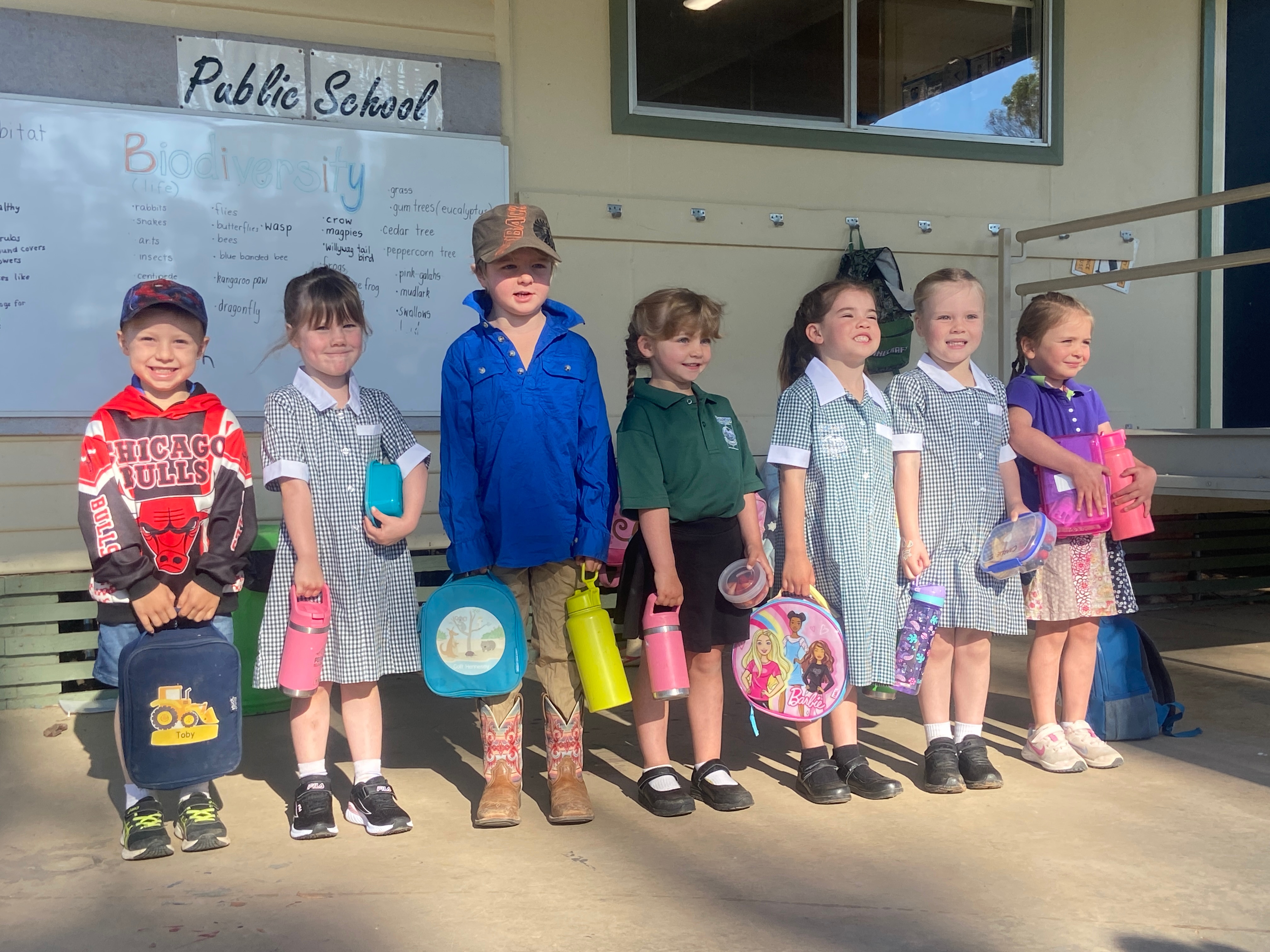 Seven children stand in a line, 4 girls are in a green school uniform while two boys and a girl wear bright clothing