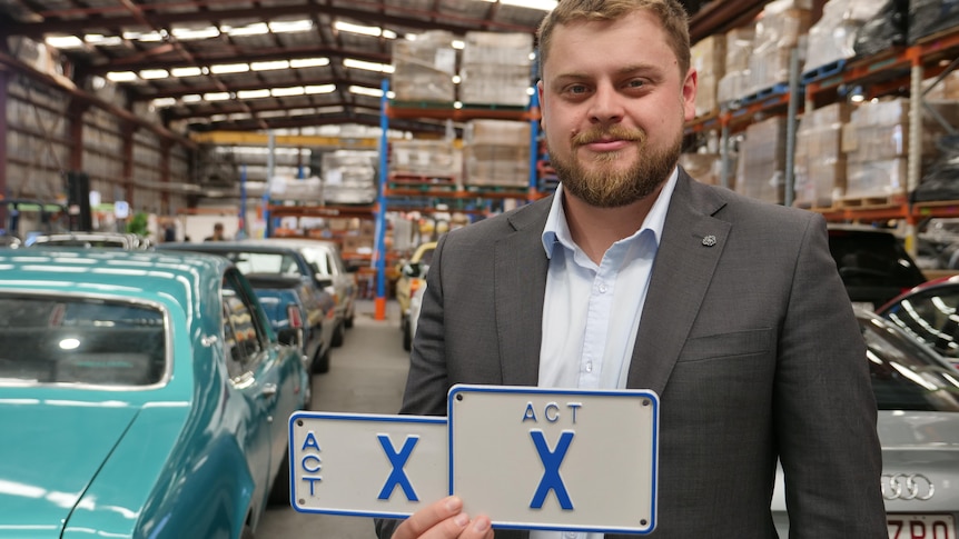 Man in suit holds a number plate that simply reads 'X'