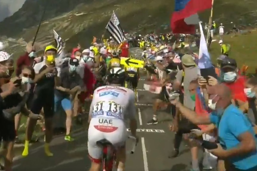 Masked Tour de France fans converge as leading riders make a large climb.
