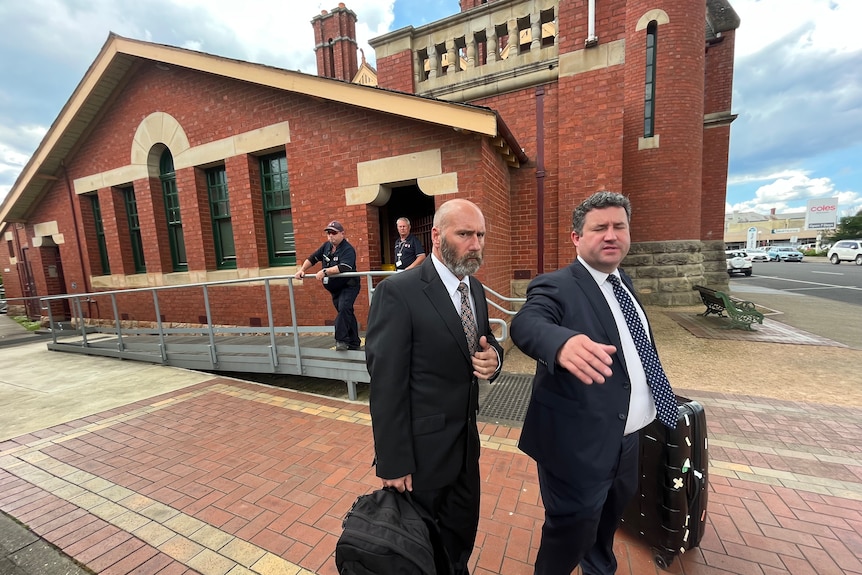Two men in suits, one with a beard, leave a small court building