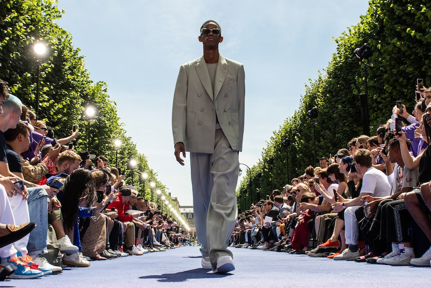 A Black male model in slouchy grey suit walks down an outdoor runway