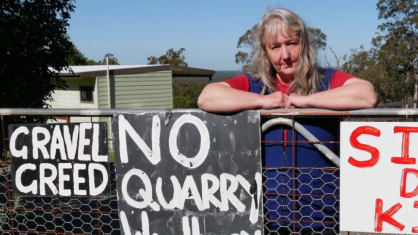 A woman with long silver hair leans against a fence with signs opposing quarry developments hanging from it.
