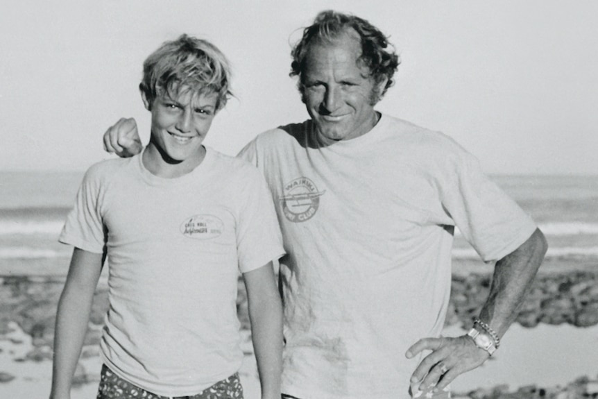 A man and a young boy smile for the camera while standing on a beach in this black and white photo.