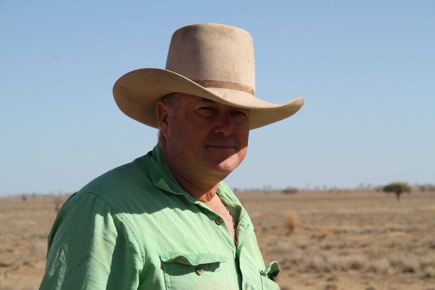 An older man in a button-up shirt, wearing a large hat, standing in a dusty paddock.