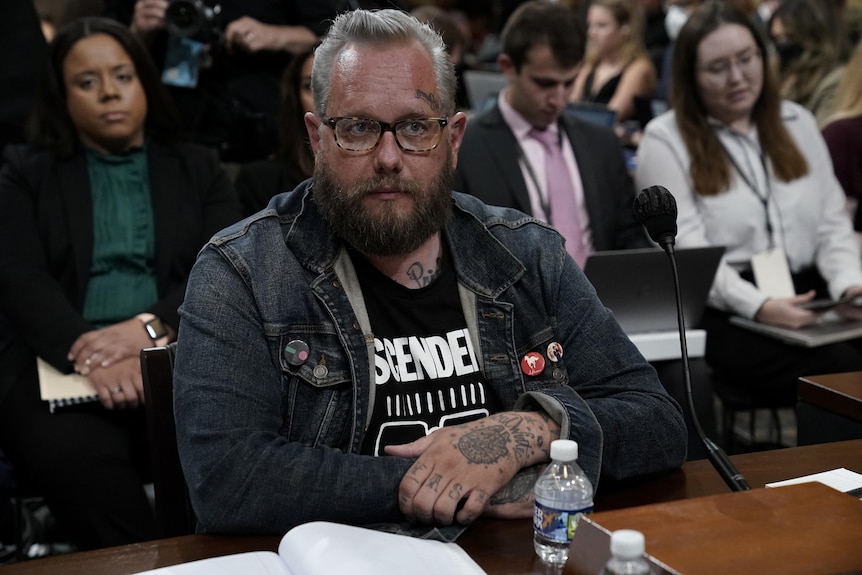 A man wearing a denim jacket with buttons on it holds his tattooed hands in front of him and rests them on a desk