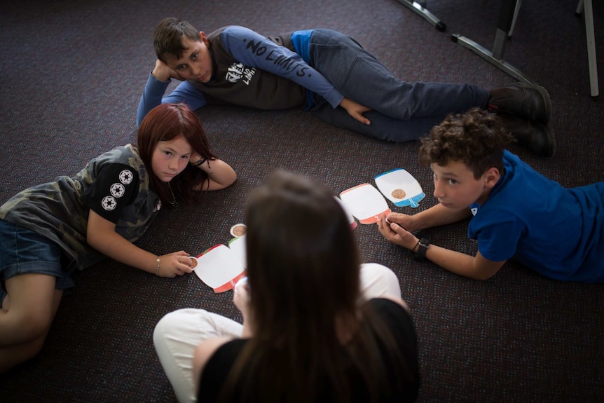 Three students lie on the carpet, eyes on teacher Anita Harding, maths problems laid out on cards on the floor between them.