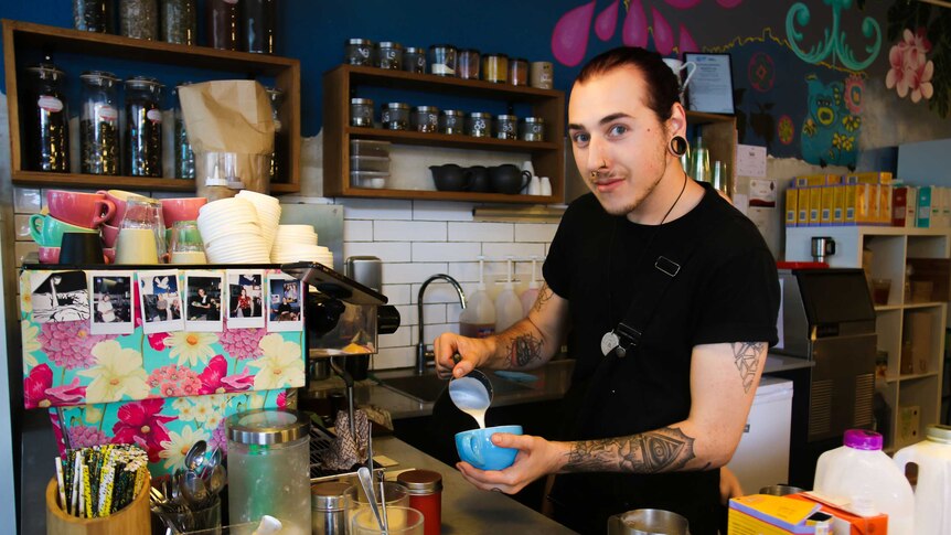 A man stands behind an coffee machine pouring milk into a blue cup of coffee.