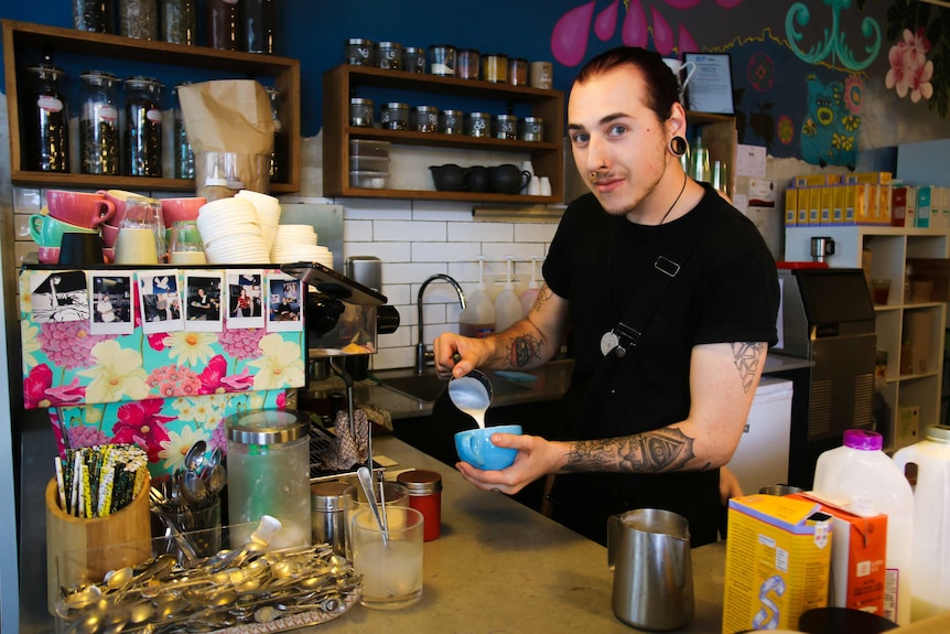A man stands behind an coffee machine pouring milk into a blue cup of coffee.