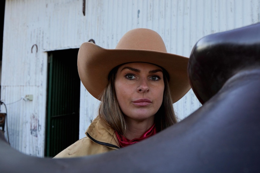 The face of a woman looking over a horse saddle while wearing a broad-brimmed hat, with a shed behind her.