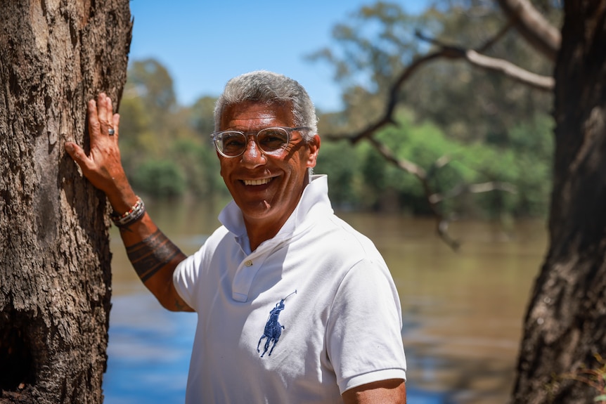 Greg James smiles wearing a ehite polo shirt, framed by gum trees, a swollen brown river visible in the background. 