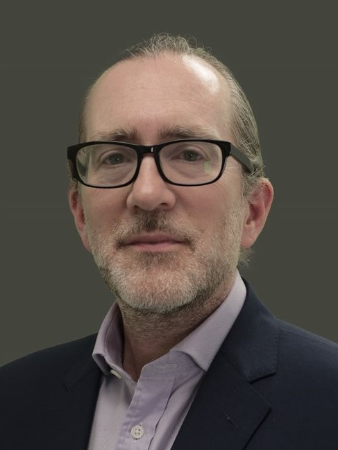 A man with black rimmed glasses, a grey beard and a blue shirt headshot
