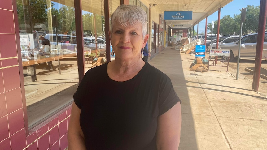 A smiling woman with grey hair stands on a street front outside a business.