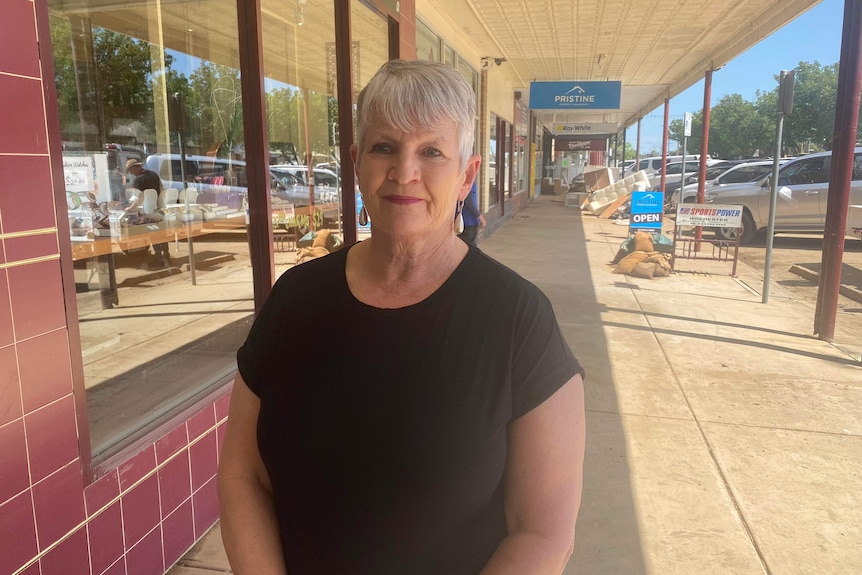 A smiling woman with grey hair stands on a street front outside a business.