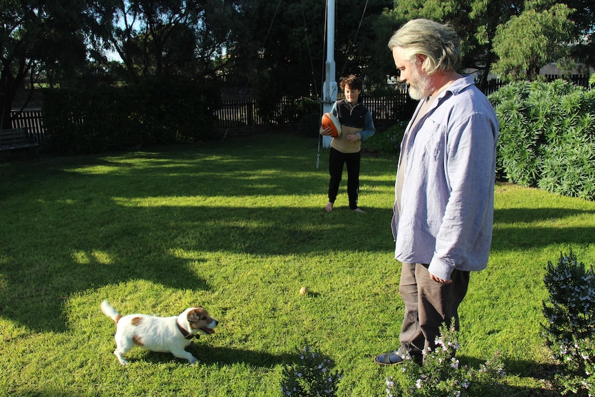 Un homme aux cheveux gris et un garçon tenant un sourire de football à un chien jack russell, le tout dans une cour gazonnée