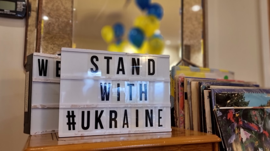 Stand with Ukraine sign sitting on a table with yellow and blue balloons in the background.